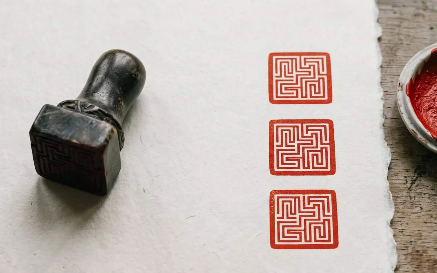 A carved stone seal on handmade paper beside three vermillion impressions, with a dish of seal paste