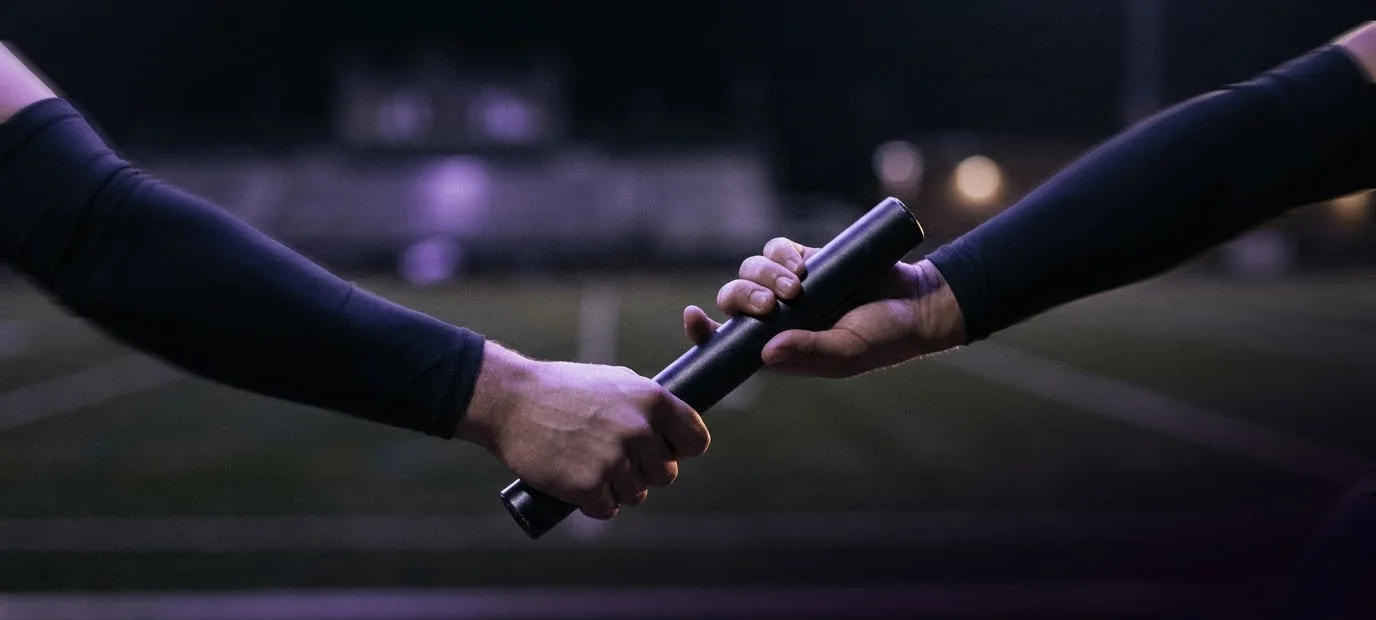 Two hands passing a relay baton on a floodlit track at night